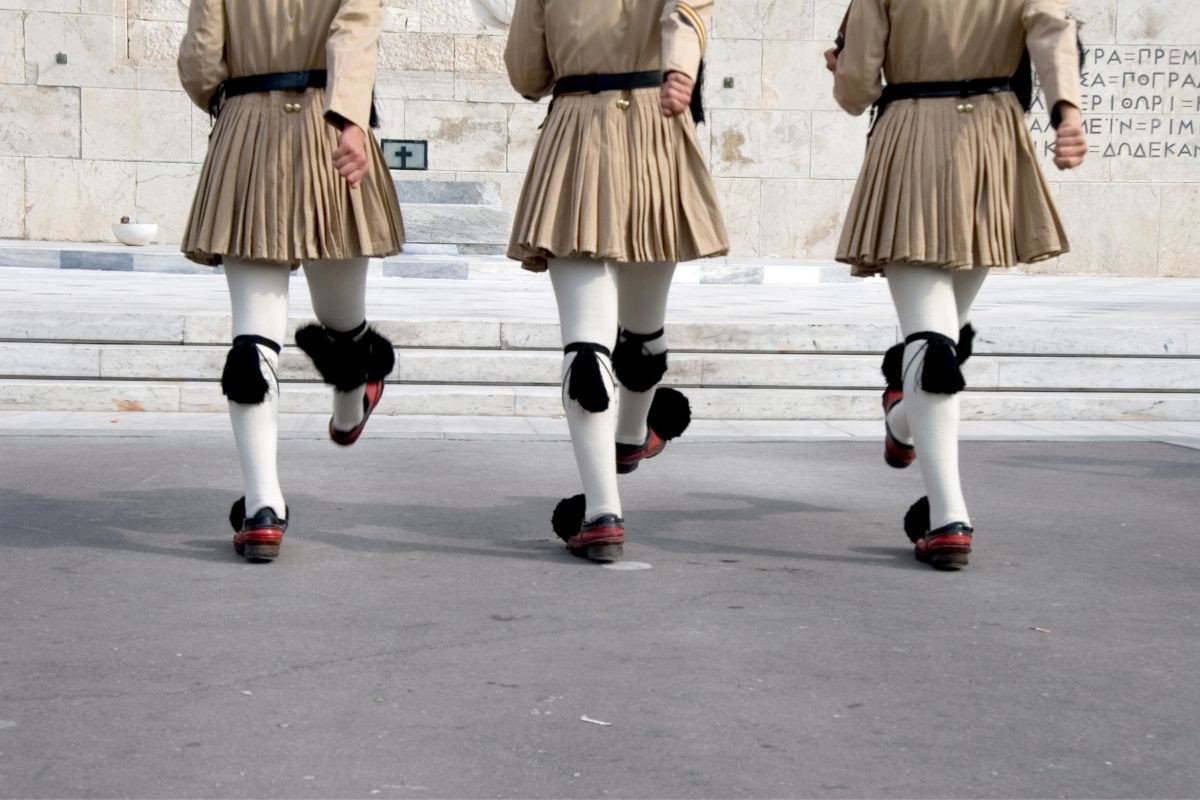 The image shows three Evzones, members of the Greek Presidential Guard, marching in traditional uniforms during the Changing of the Guard ceremony.